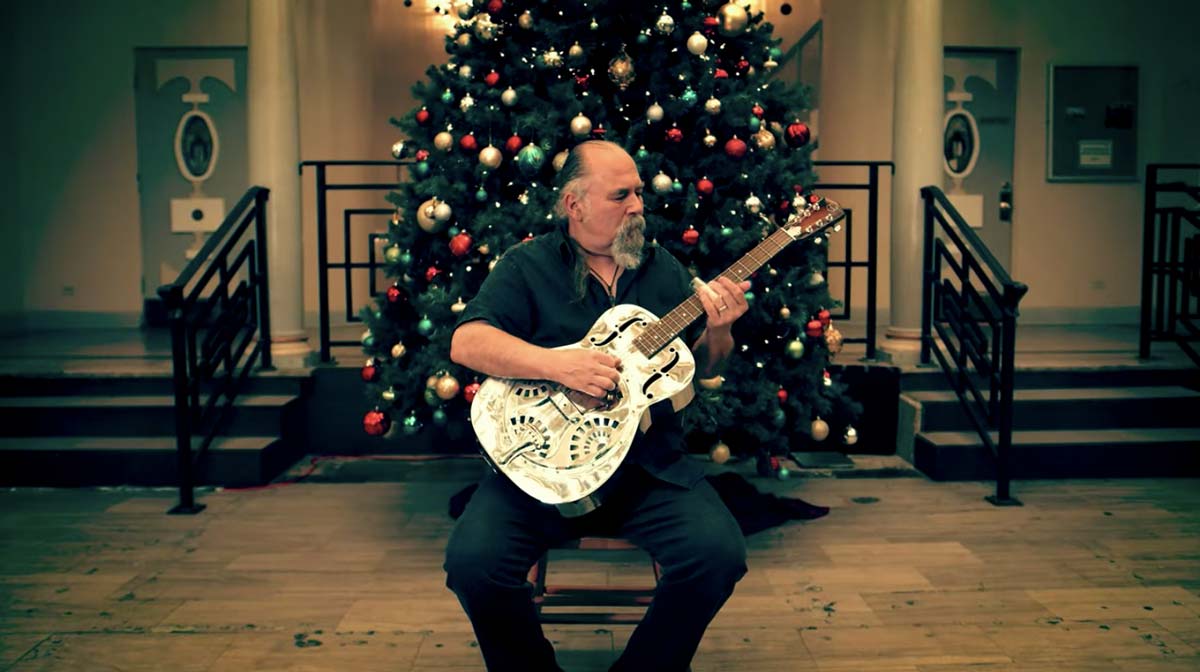 Glenn Kaiser playing a guitar in front of a large Christmas tree in a building lobby